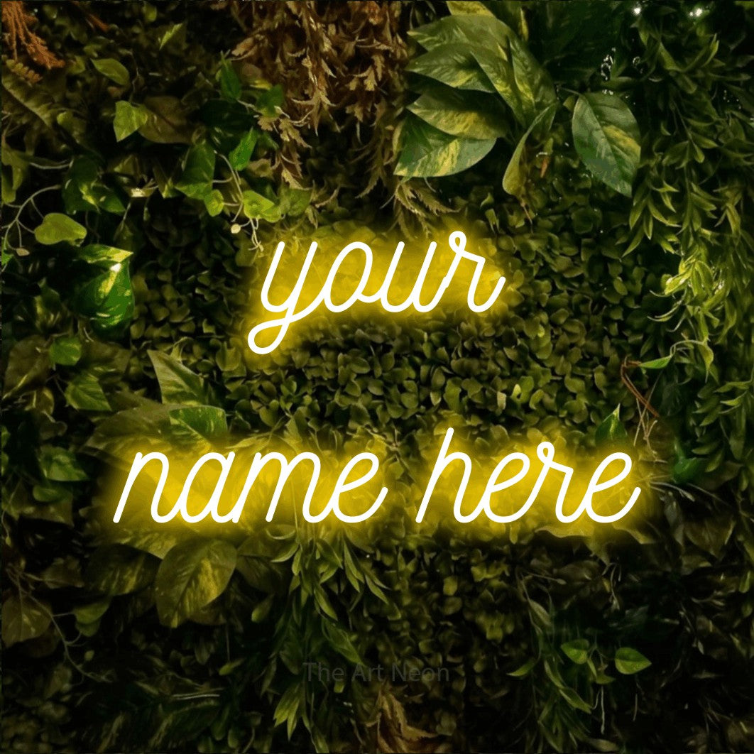 Yellow script-style LED neon sign reading "Your Name Here" against a textured moss and foliage wall for office décor