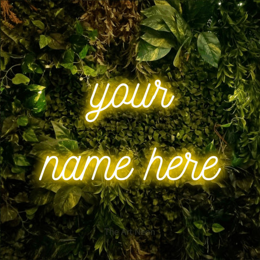 Yellow script-style LED neon sign reading "Your Name Here" against a textured moss and foliage wall for office décor