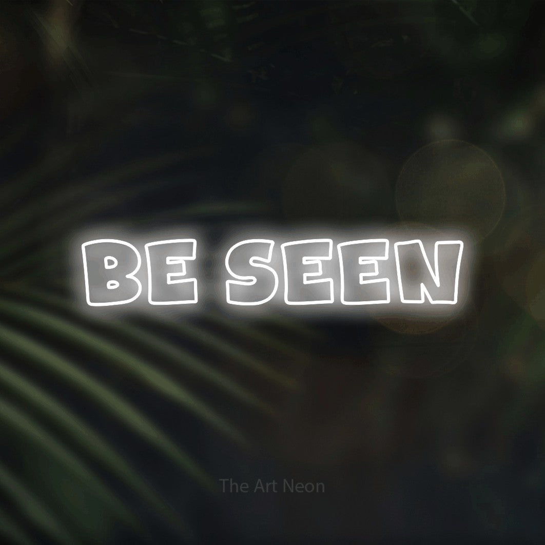 Cool white outline-style LED neon sign reading "Be Seen" against dark blurred palm leaves for office décor