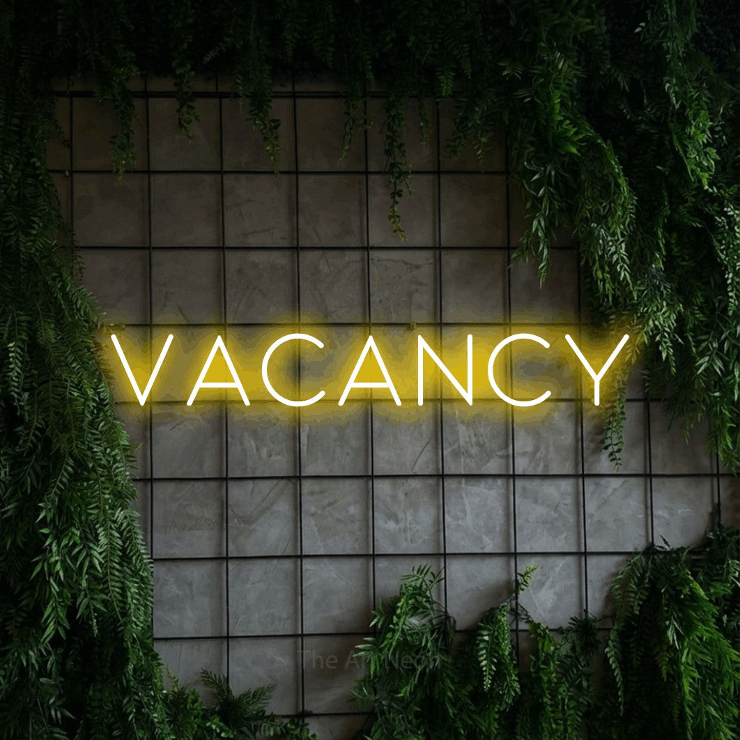 Yellow LED neon sign reading "Vacancy" against a green grid wall with foliage for hotel décor