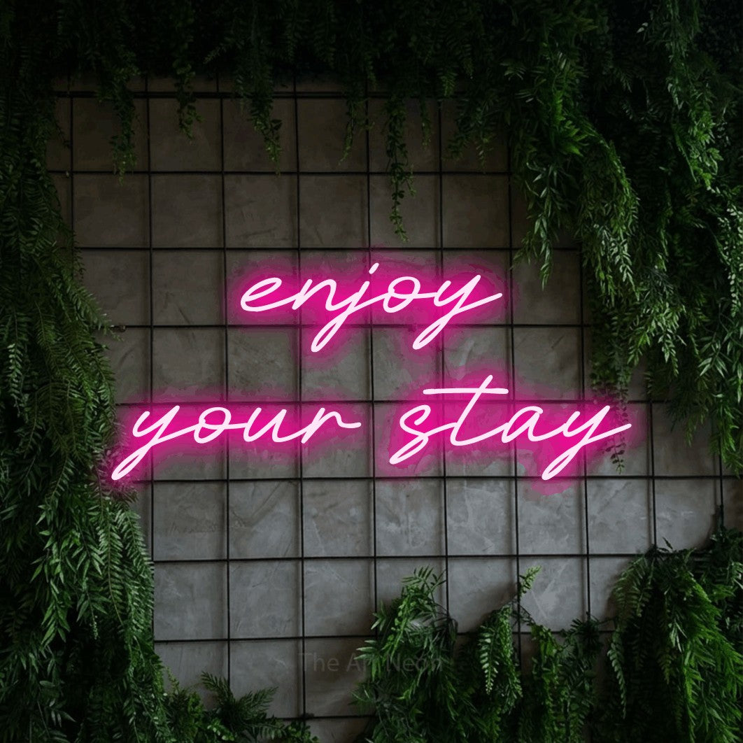Hot pink LED neon sign reading "Enjoy Your Stay" against a green grid wall with foliage for hotel décor