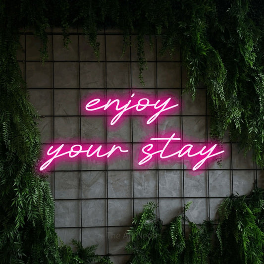 Hot pink LED neon sign reading "Enjoy Your Stay" against a green grid wall with foliage for hotel décor