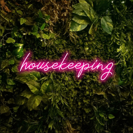Hot pink script-style LED neon sign reading "Housekeeping" against a textured moss and foliage wall for hotel décor