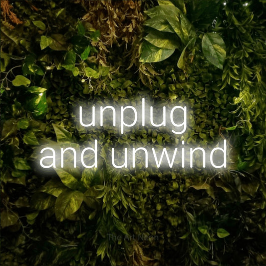 Cool white LED neon sign reading "Unplug and Unwind" against a textured moss and foliage wall for hotel décor