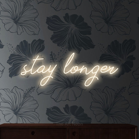 Warm white script-style LED neon sign reading "Stay Longer" against grey floral wallpaper for hotel décor