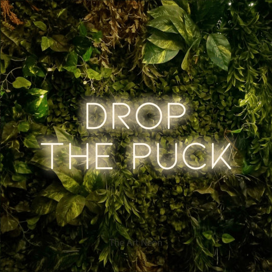 Enseigne néon à DEL à lumière blanche chaude, affichant « Drop the Puck » sur un mur texturé de mousse et de feuillage