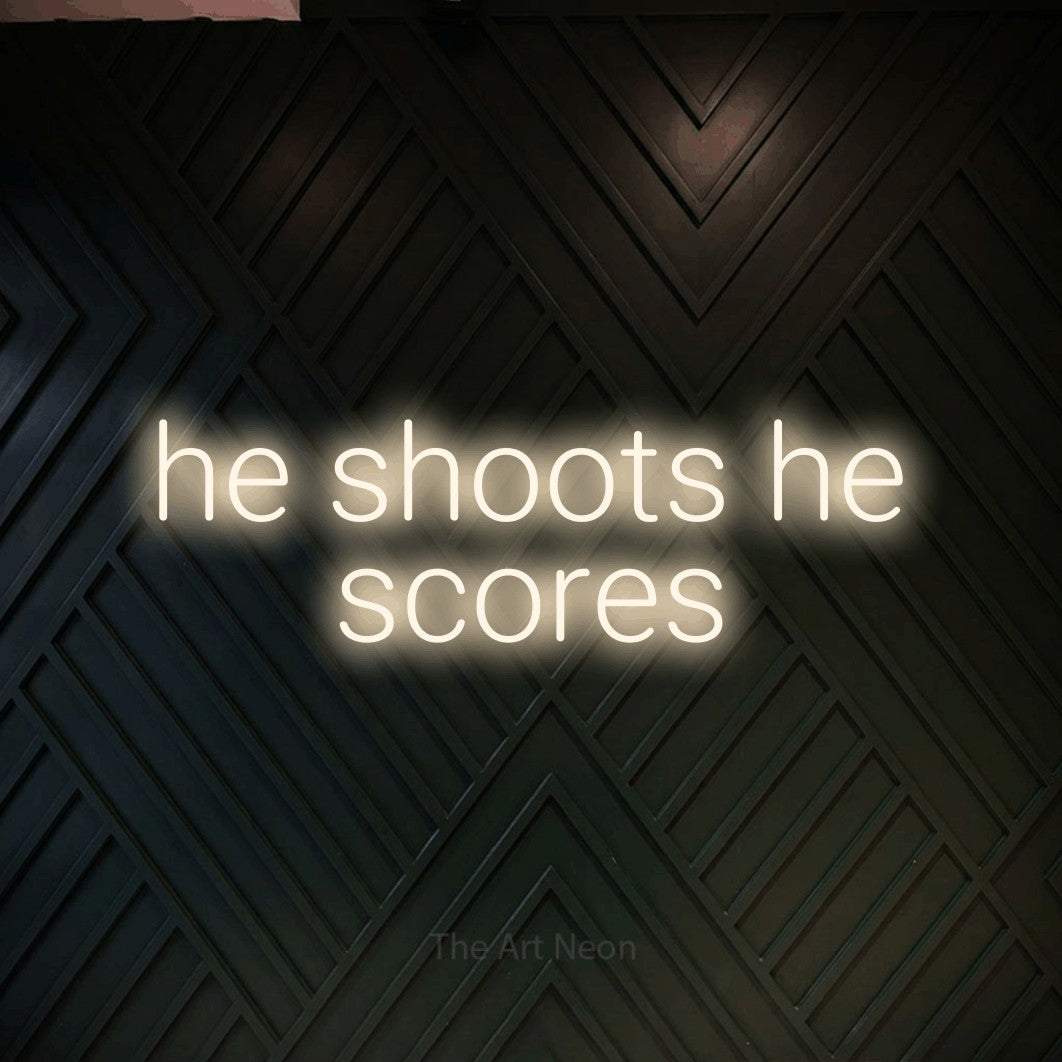 Warm white LED neon sign reading "He Shoots He Scores" against a dark geometric wood wall
