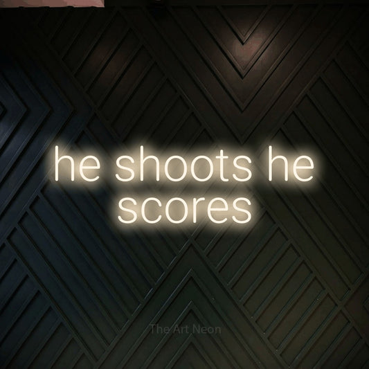 Enseigne néon à DEL à lumière blanche chaude, affichant « He Shoots He Scores » sur un mur géométrique en bois foncé