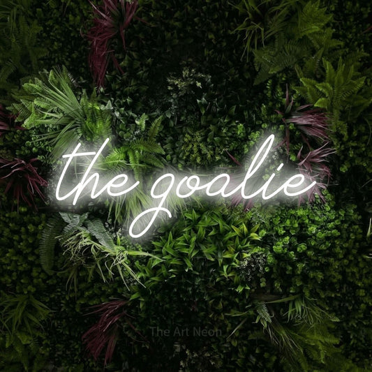 Cool white script-style LED neon sign reading "The Goalie" against a lush garden wall with pink flowers