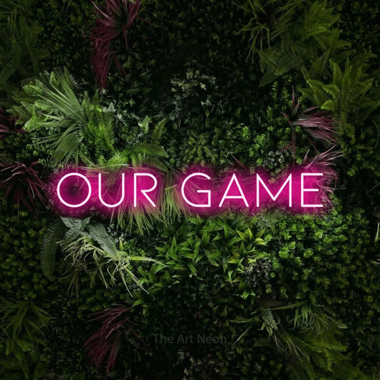 Hot pink LED neon sign reading "Our Game" against a lush garden wall with pink flowers