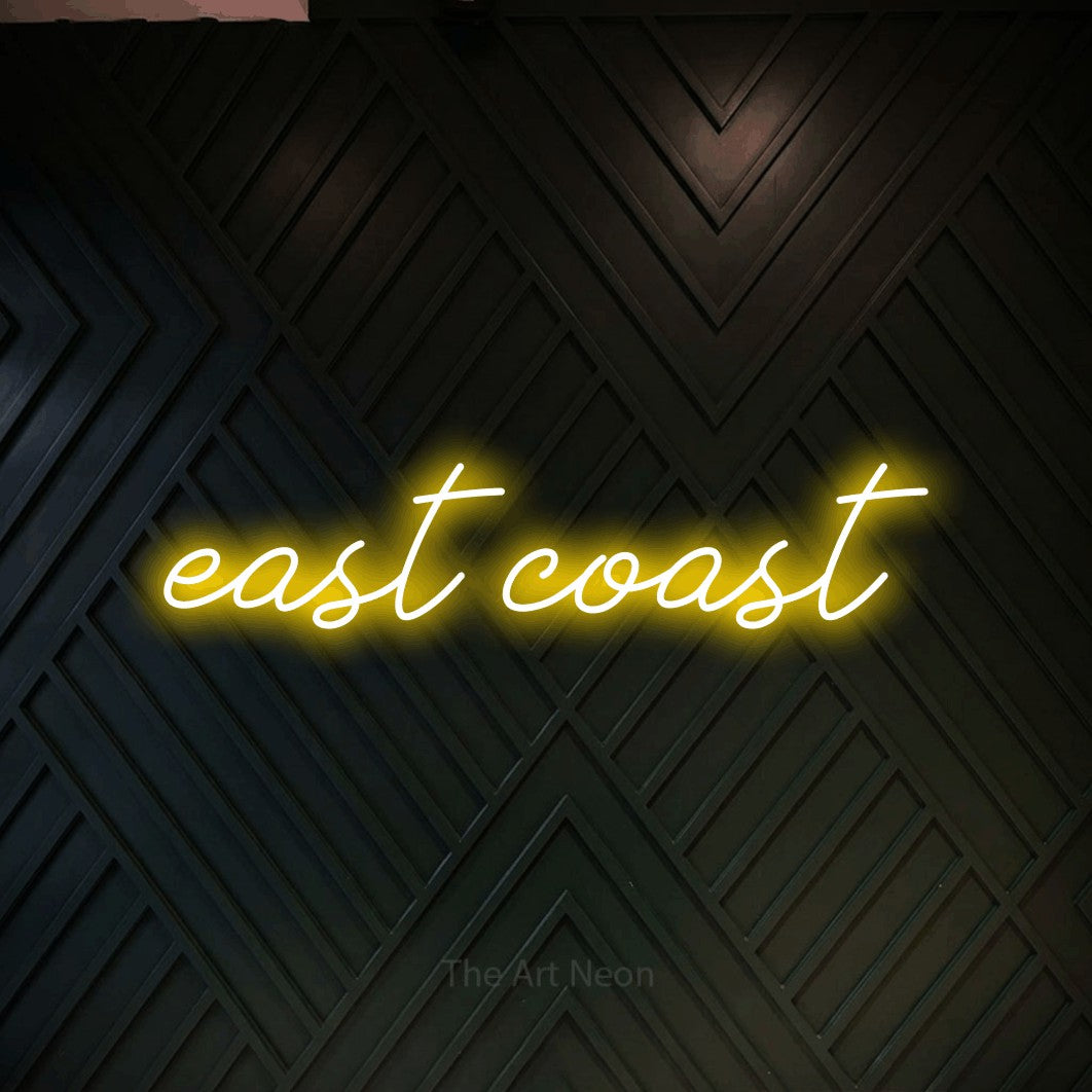 Yellow script-style LED neon sign reading "East Coast" against a dark geometric wood wall