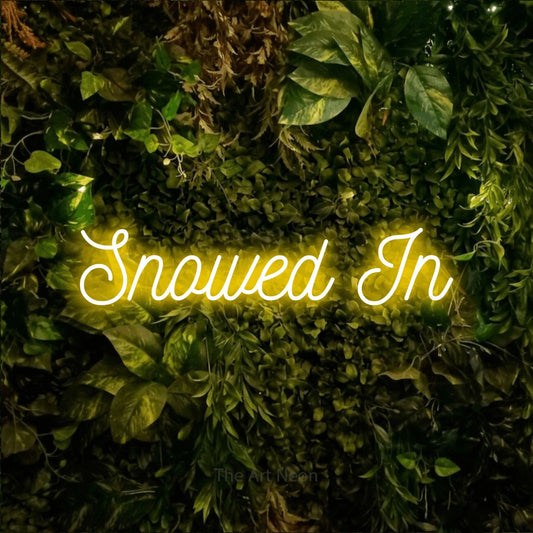 Yellow script-style LED neon sign reading "Snowed In" against a textured moss and foliage wall