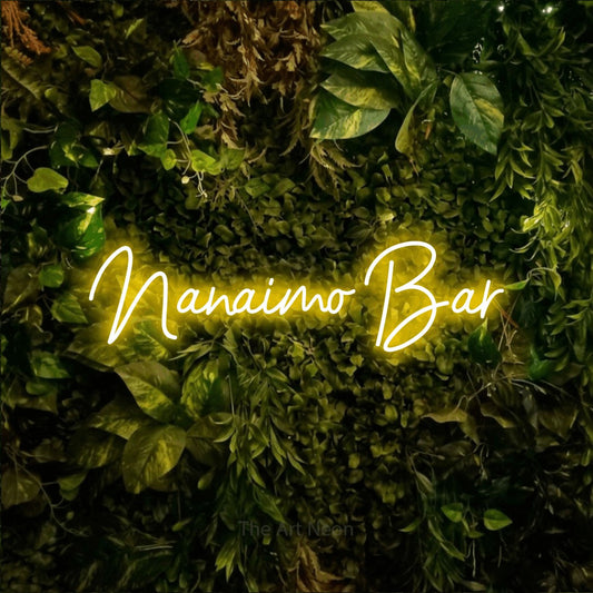 Enseigne néon à DEL à lumière jaune, de style manuscrit, affichant « Nanaimo Bar » sur un mur texturé de mousse et de feuillage