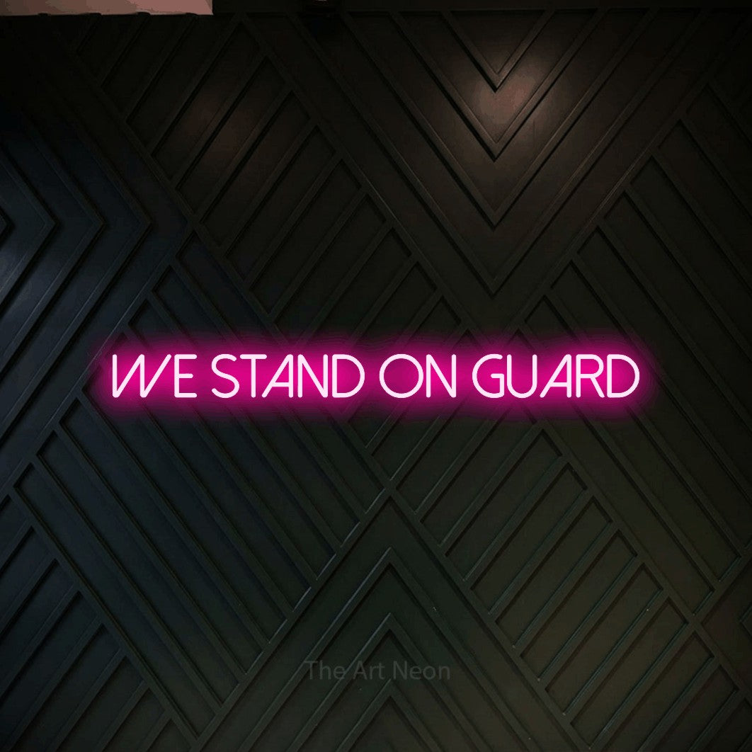 Hot pink LED neon sign reading "We Stand on Guard" against a dark geometric wood wall