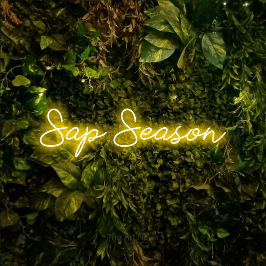 Yellow script-style LED neon sign reading "Sap Season" against a textured moss and foliage wall