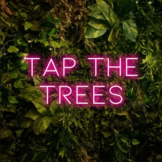 Hot pink LED neon sign reading "Tap the Trees" against a textured moss and foliage wall