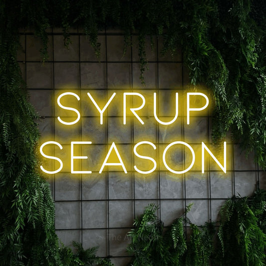 Yellow LED neon sign reading "Syrup Season" against a green grid wall with foliage