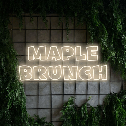 Warm white outline-style LED neon sign reading "Maple Brunch" against a green grid wall with foliage