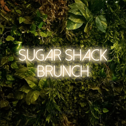 Warm white LED neon sign reading "Sugar Shack Brunch" against a textured moss and foliage wall