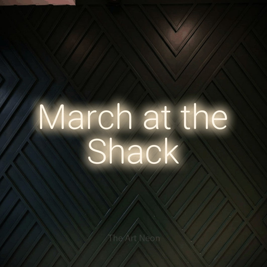 Warm white LED neon sign reading "March at the Shack" against a dark geometric wood wall