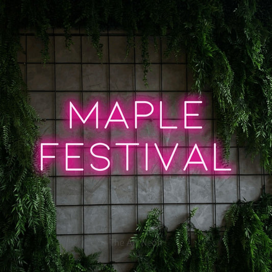 Hot pink LED neon sign reading "Maple Festival" against a green grid wall with foliage
