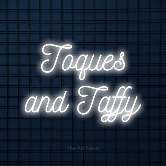 Cool white script-style LED neon sign reading "Toques and Taffy" against a dark blue tiled wall