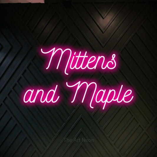Hot pink script-style LED neon sign reading "Mittens and Maple" against a dark geometric wood wall