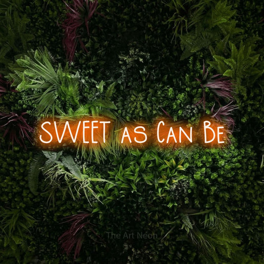 Orange LED neon sign reading "Sweet as Can Be" against a lush garden wall with pink flowers