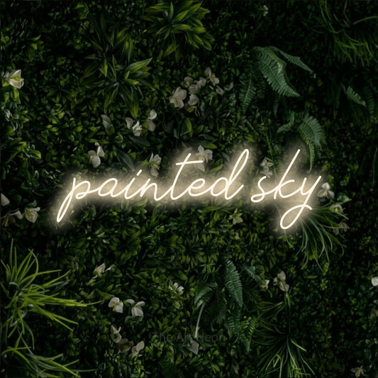 Warm white script-style LED neon sign reading "Painted Sky" against a leafy green wall
