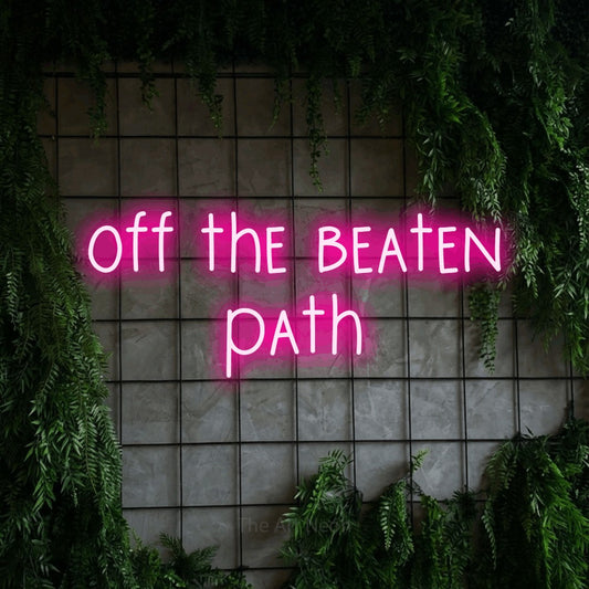 Hot pink LED neon sign reading "Off the Beaten Path" against a green grid wall with foliage