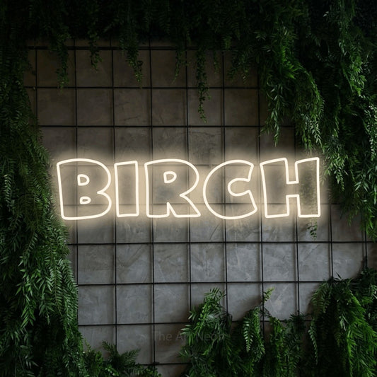Warm white outline-style LED neon sign reading "Birch" against a green grid wall with foliage
