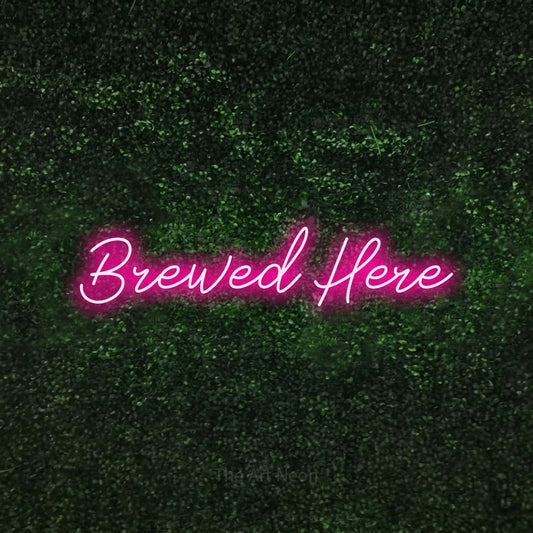 Hot pink script-style LED neon sign reading "Brewed Here" against a textured green moss wall