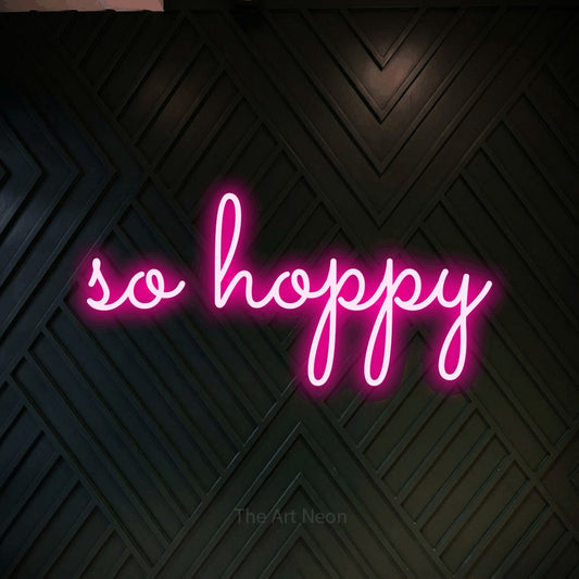 Hot pink LED neon sign reading "So Hoppy" against a dark geometric wood wall