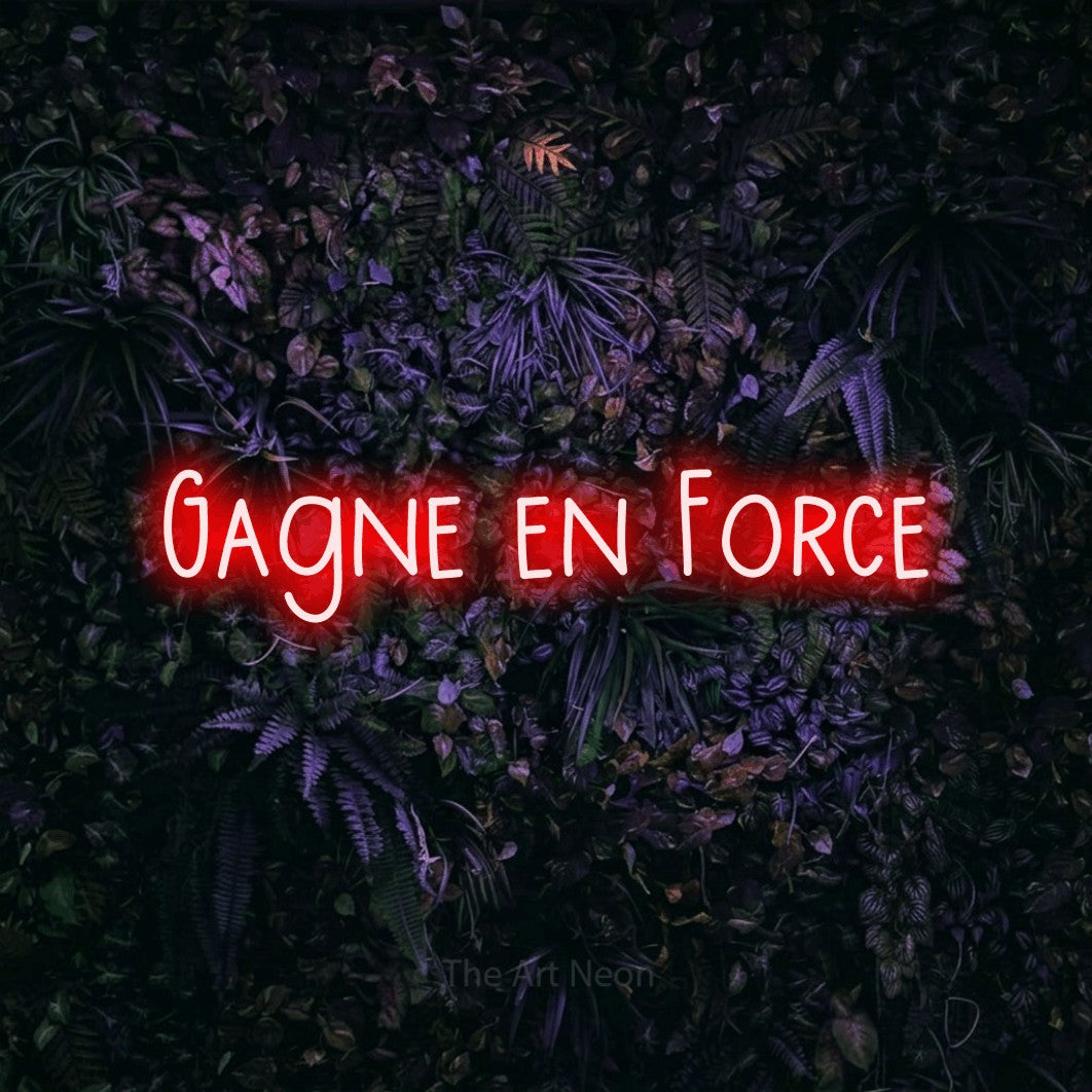 “Gagne en force” LED neon sign in red, with narrow hand-drawn lettering, shown against a dark wall with purple flowers.