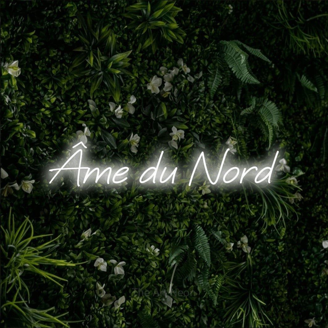 “Âme du nord” LED neon sign in cool white, with slanted cursive lettering, shown against a dense green plant wall.