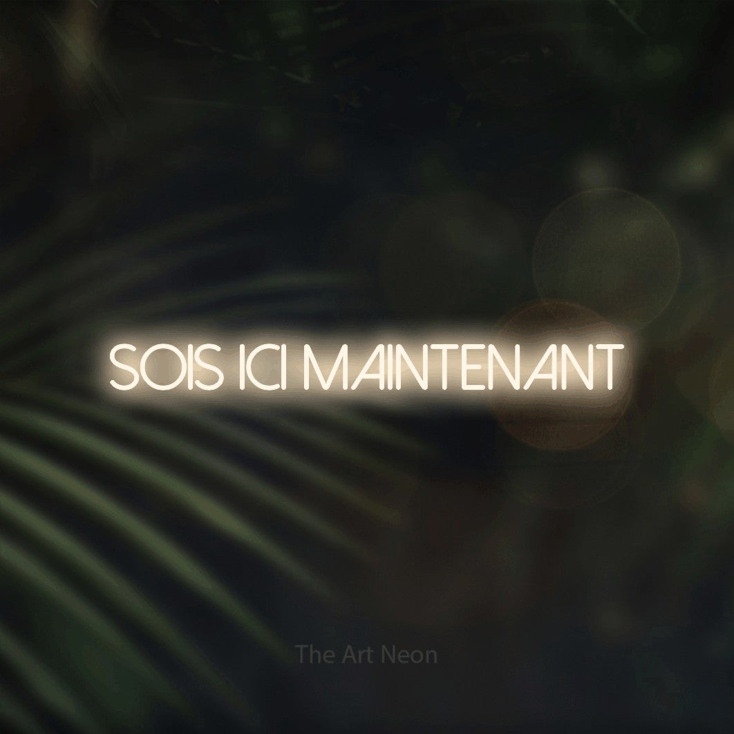“Sois ici maintenant” LED neon sign in warm white, with thin sans-serif lettering, shown against a blurred palm and bokeh-light backdrop.