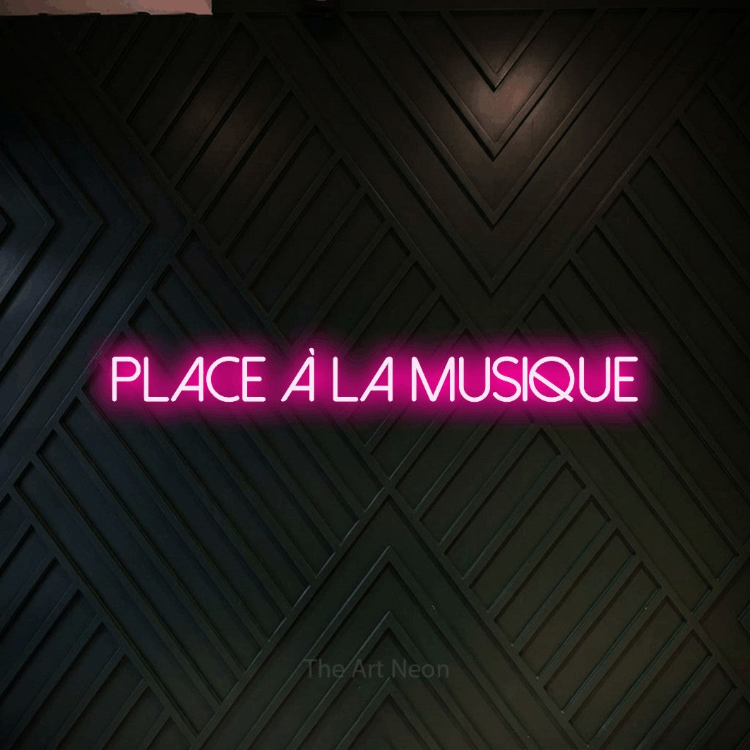 “Place à la musique” LED neon sign in hot pink, with thin sans-serif lettering, shown against a black chevron-patterned wood wall.