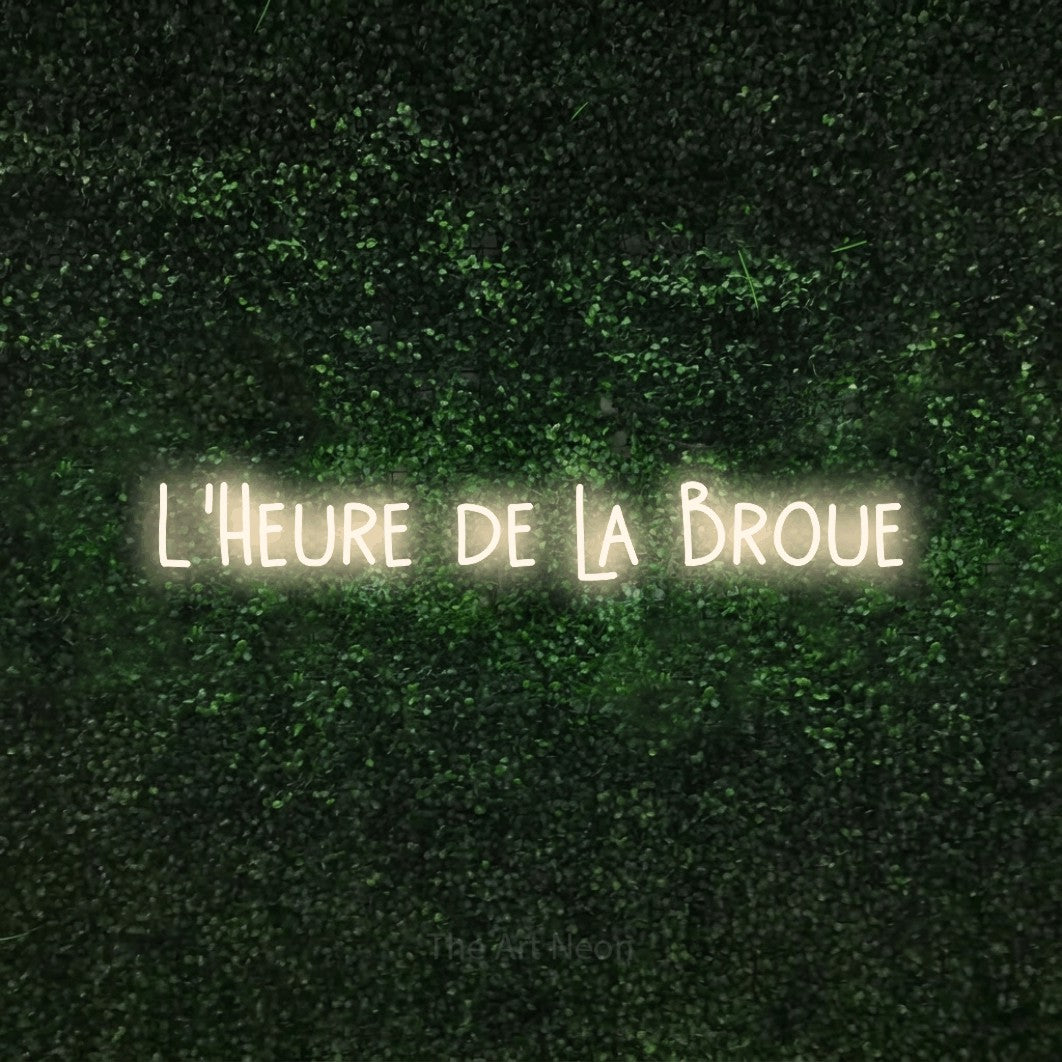 “L'heure de la broue” LED neon sign in warm white, with narrow hand-drawn lettering, shown against a textured green hedge.