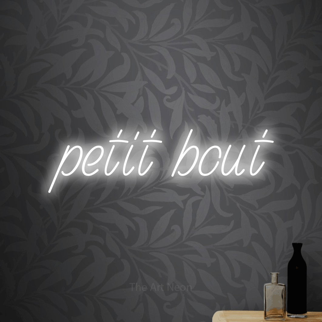 “Petit bout” LED neon sign in cool white, with slanted handwritten lettering, shown against grey botanical wallpaper.