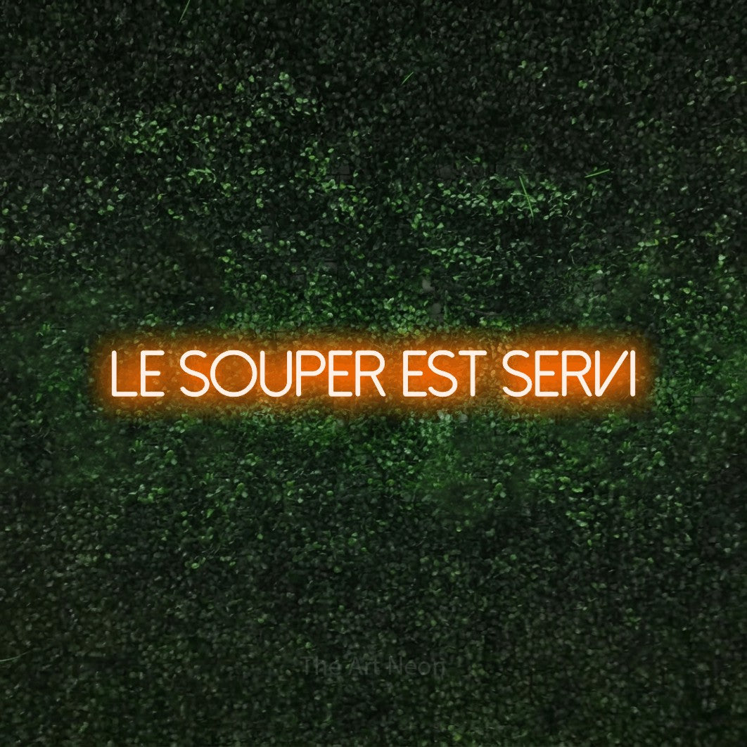 “Le souper est servi” LED neon sign in orange, with thin sans-serif lettering, shown against a textured green hedge.