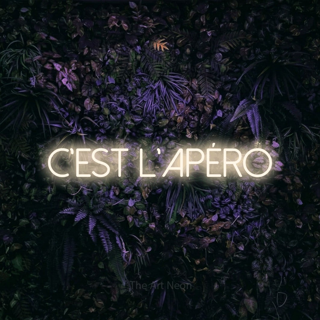 “C'est l'apéro” LED neon sign in warm white, with thin sans-serif lettering, shown against a dark wall with purple flowers.