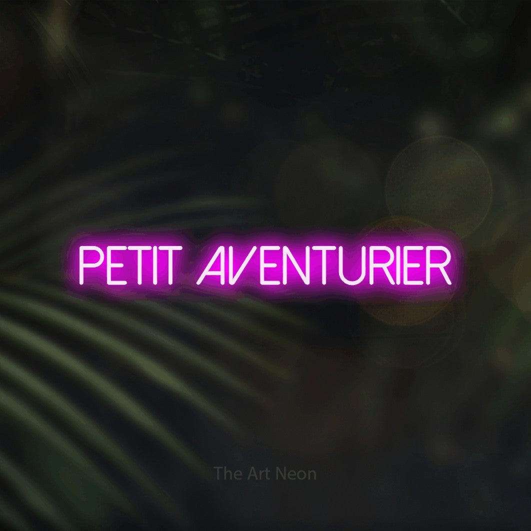 “Petit aventurier” LED neon sign in pink, with thin sans-serif lettering, shown against a blurred palm and bokeh-light backdrop.