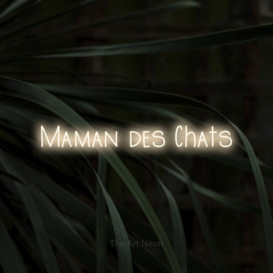 “Maman des chats” LED neon sign in warm white, with narrow hand-drawn lettering, shown against a close-up of dark foliage.