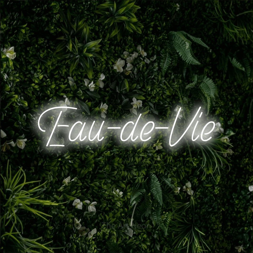 “Eau-de-vie” LED neon sign in cool white, with slanted handwritten lettering, shown against a dense green plant wall.