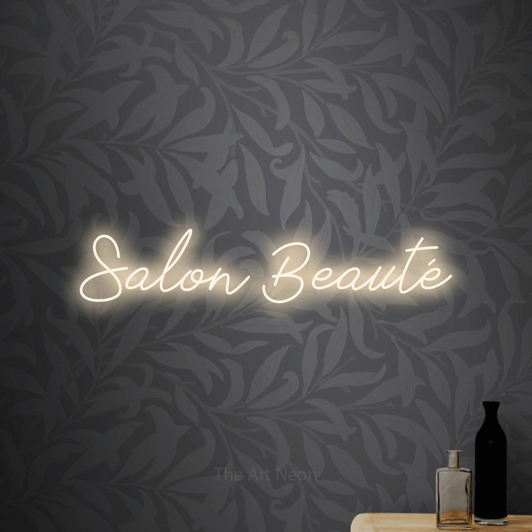 Composition en néon LED « Salon beauté » en blanc chaud, en lettrage cursif, mise en valeur sur un papier peint botanique gris, pour un salon de beauté.