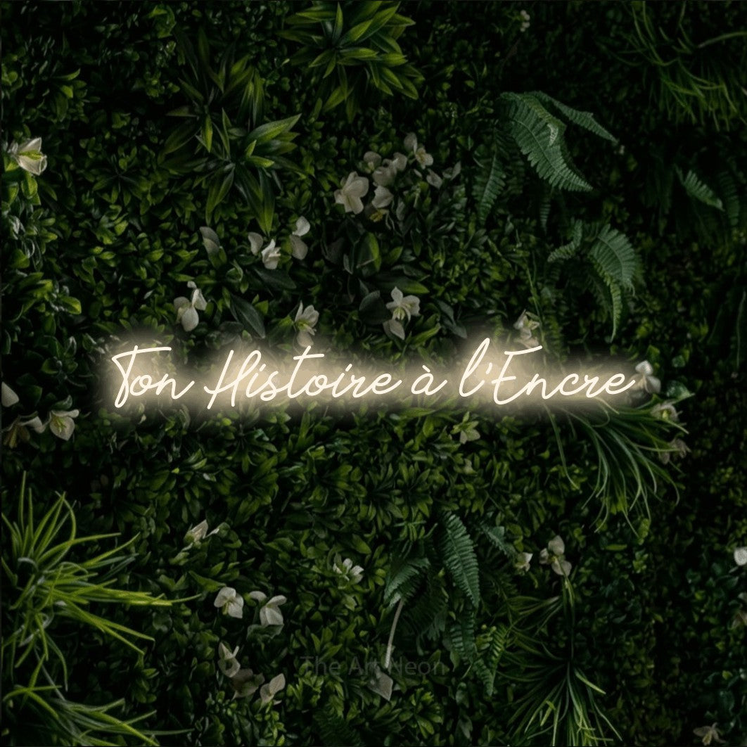 “Ton histoire à l'encre” LED neon sign in warm white, with cursive lettering, shown against a dense green plant wall.