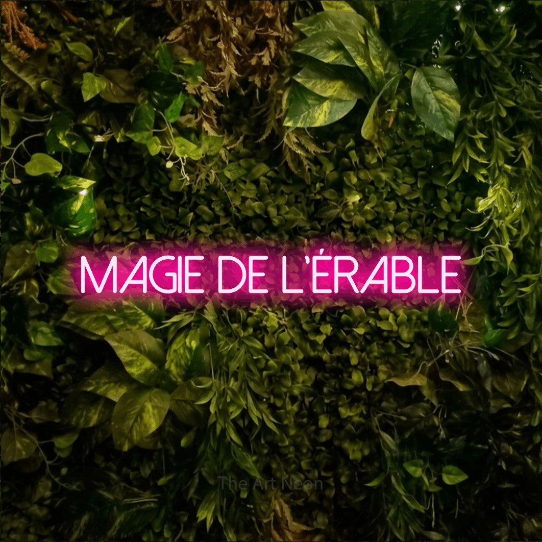 Aperçu d’une enseigne néon LED « Magie de l'érable » en rose vif, en lettres fines sans empattement, photographiée sur un feuillage vert et jaune vif, pour un décor canadien.