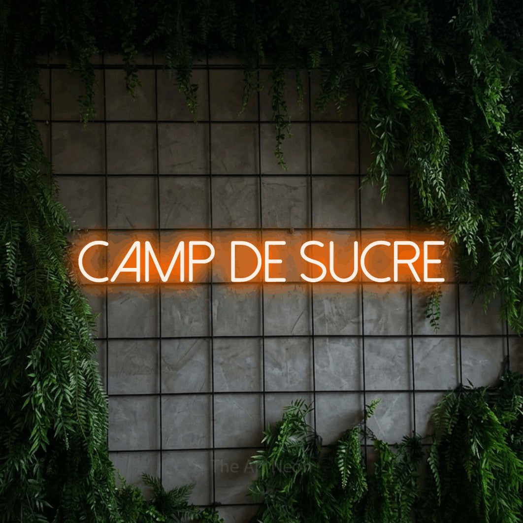 “Camp de sucre” LED neon sign in orange, with thin sans-serif lettering, shown against an ivy-covered concrete grid.