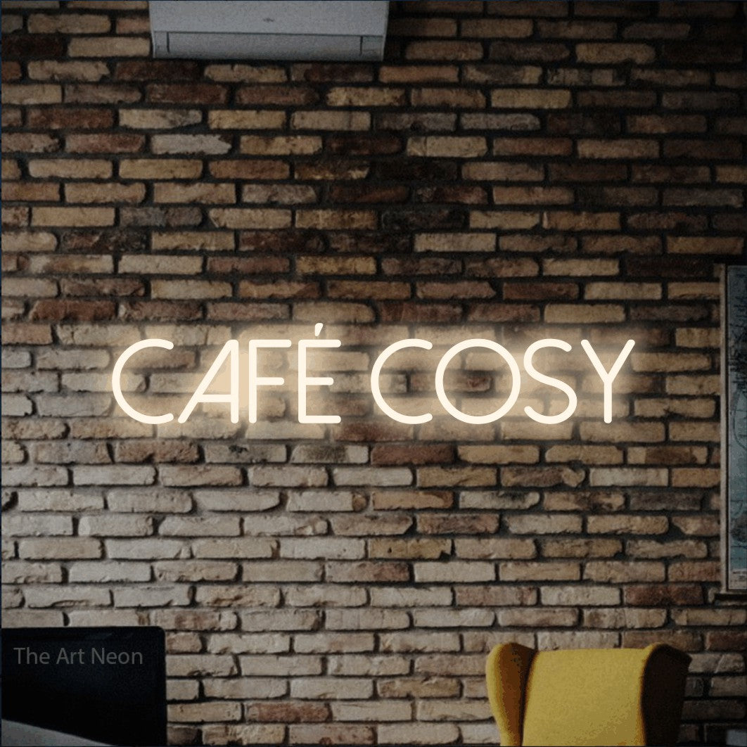 Composition en néon LED « Café cosy » en blanc chaud, en lettres fines sans empattement, mise en valeur sur un mur de briques aux tons chauds, pour un café.