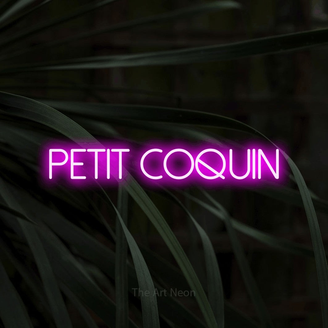 “Petit coquin” LED neon sign in pink, with thin sans-serif lettering, shown against a close-up of dark foliage.